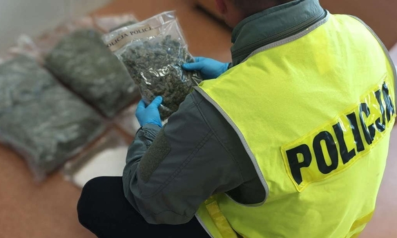 policjant trzyma foliowy woreczek z suszem roślinnym, w tle na podłodze leżą foliowe woreczki z suszem roślinnym