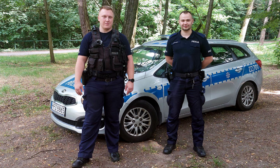 dwaj policjanci stoją przed radiowozem policyjnym, w tle drzewa