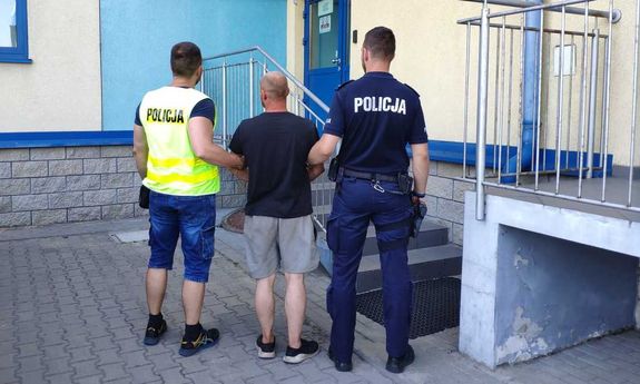 dwaj  policjanci prowadzą zatrzymanego. Jeden z policjantów ma na sobie kamizelkę z napisem Policja, drugi jest w policyjnym mundurze