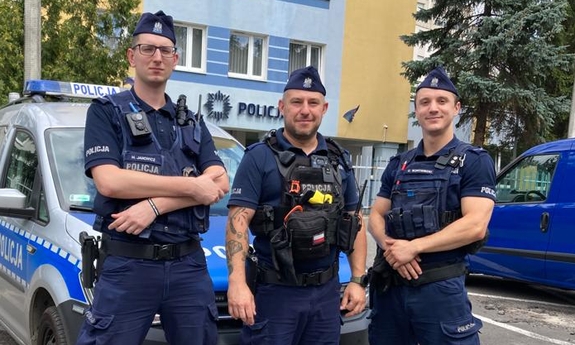 trzej policjanci stoją przy radiowozie policyjnym na parkingu przed budynkiem komendy Policji