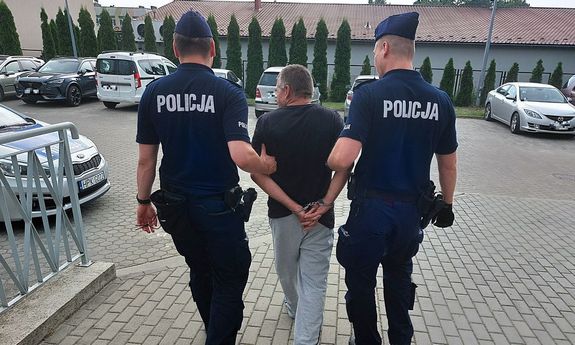Zatrzymany jest prowadzony przez dwóch umundurowanych funkcjonariuszy. Mężczyzna ma kajdanki założone na rękach trzymanych z tyłu