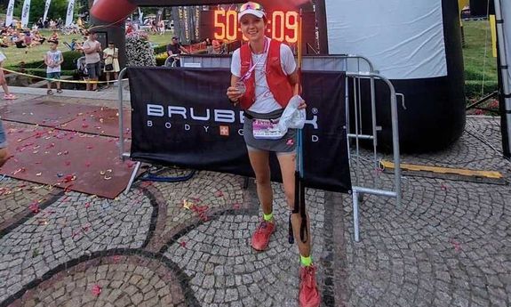 Na pierwszym planie stojąca kobieta w stroju sportowym trzymająca medal. Za nią widoczna dmuchana meta oraz zegar cyfrowy