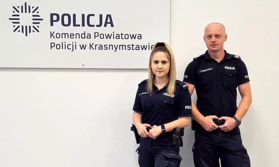 policjant o policjantka pozują do zdjęcia, na białej ścianie napis Policja Komenda Powiatowa Policji w Krasnymstawie