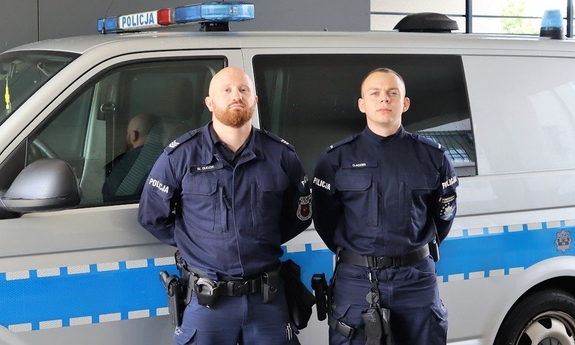 na tle policyjnego radiowozu stoją dwaj umundurowani policjanci