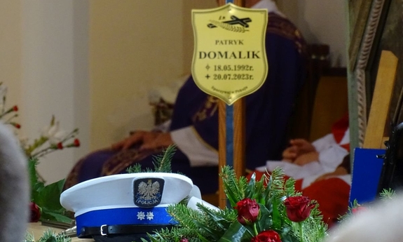 czapka policyjna leżąca ma trumnie w kościele, w tle na krzyżu tabliczka z imieniem i nazwiskiem zmarłego policjanta oraz data jego urodzin i śmierci