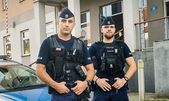 dwaj umundurowani policjanci stojący przy radiowozie, w tle budynek komendy Policji