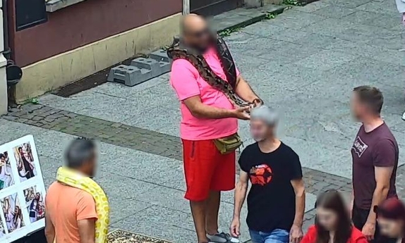 Mężczyzna stoi na ulicy z wężem na szyi, ulicą idą ludzie