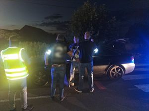 Zdjęcie wykonane nocą. Przy samochodzie tyłem stoi zatrzymany mężczyzna, po jego obu stronach policjanci w policyjnych opaskach na ramieniu. Pol lewej stronie funkcjonariusz w kamizelce z napisem policja