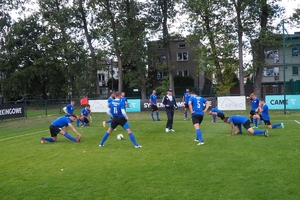 mężczyźni przebrani w stroje sportowe rozgrzewają się przed meczem na boisku, na środku stoi trener drużyny piłkarskiej