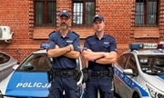 Dwaj umundurowani policjanci stojący na tle zaparkowanych radiowozów i budynku z cegły