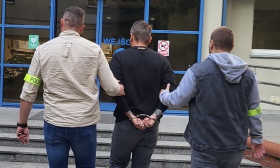 dwaj nieumundurowani policjanci prowadzą zatrzymanego mężczyznę zakutego w kajdanki w stronę wejścia do budynku