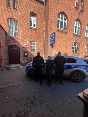 dwaj policjanci i policjantka stoją przy radiowozie policyjnym, w tle budynek komendy Policji