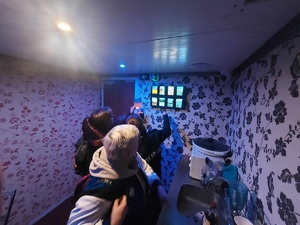 Wnętrze escapetrucka stylizowane na kuchnię, po prawej stronie zlew z naczyniami, na ścianie osiem małych ekranów dotykowych. Policjantka i dwie osoby próbują rozwiązać zagadki.
