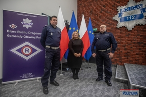 dwaj policjanci i kobieta stojący na tle ściany i flag Polski i Unii Europejskiej