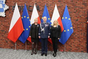 Odznaczony medalem Komendant Główny Policji stoi na tle flag Polski i Unii Europejskiej. Obok niego stoją dwie kobiety ubrane na czarno