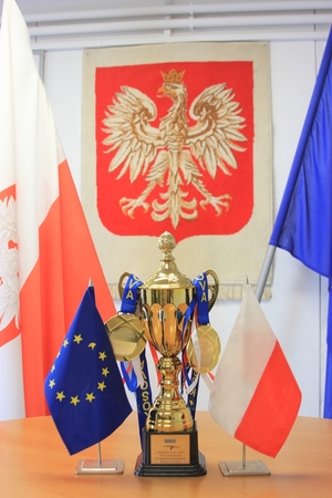 na stole stoi puchar, po jego bokach miniaturki flag Polski i Unii europejskiej, w tle na ścianie wisi godło Polski, a po bokach flagi Polski i Unii Europejskiej