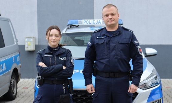 Na tle szarego budynku i oznakowanego radiowozu stoi dwoje policjantów