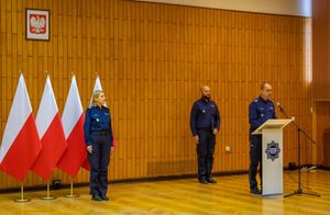 przy mównicy stoi policjant przemawia do mikrofonu, za nią przy ścianie po lewej i prawej stronie stoi dwoje policjantów, za policjantką z lewej strony stoją cztery flagi Polski w stojakach