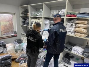 Funkcjonariusze przeszukują pomieszczenie, na regałach leżą ubrania zapakowane w foliowe worki
