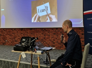 policjant siedzi przy stoliku, na którym stoi laptop, trzyma w prawej dłoni mikrofon, na ekranie wiszącym na ścianie wyświetlana jest prezentacja