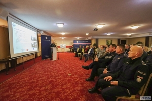 policjanci siedzą w rzędach na krzesłach słuchając policjanta, który stoi przy mównicy, na ekranie zawieszonym na ścianie wyświetlana jest prezentacja
