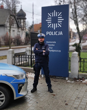 Policjant stoi przy pylonie informacyjnym z napisem Komenda Miejska Policji w Bydgoszczy