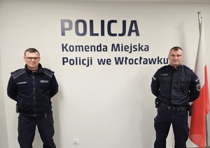 Na zdjęciu dwóch umundurowanych policjantów stoi na tle ściany z granatowym napisem Policja, Komenda Miejska Policji we Włocławku. Po prawej stronie wisi biało-czerwona flaga Polski