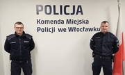 Na zdjęciu dwóch umundurowanych policjantów stoi na tle ściany z granatowym napisem Policja, Komenda Miejska Policji we Włocławku. Po prawej stronie wisi biało-czerwona flaga Polski