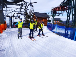 Czworo narciarzy w kamizelkach odblaskowych zjeżdża ze stoku narciarskiego.