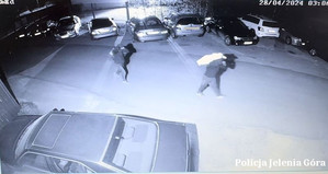Czarno-biały kadr na tle zaparkowanych samochodów  widoczni sprawcy kradzieży,  którzy idą z workami na plecach