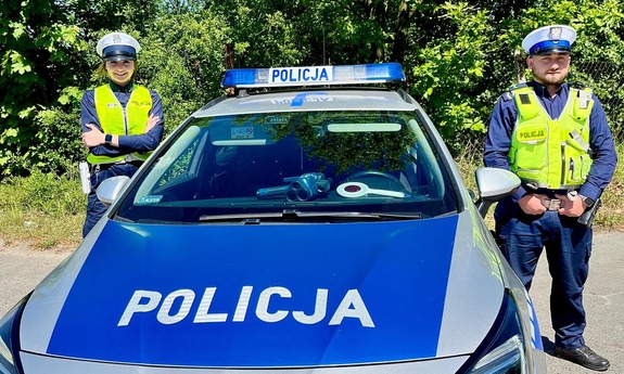 na zdjęciu policjantka i policjant stoją przy radiowozie