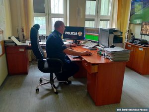 dyżurny policjant siedzi przed komputerem, za biurkiem. Widok z boku