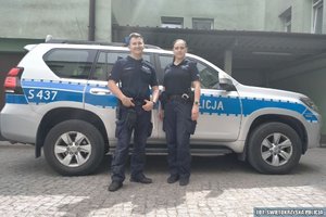 dwoje umundurowanych policjantów stoi przy oznakowanym radiowozie