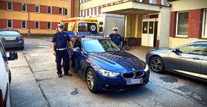 dwaj umundurowani policjanci przy nieoznakowanym radiowozie zaparkowanym przed szpitalem. Z tyłu widać karetkę pogotowia
