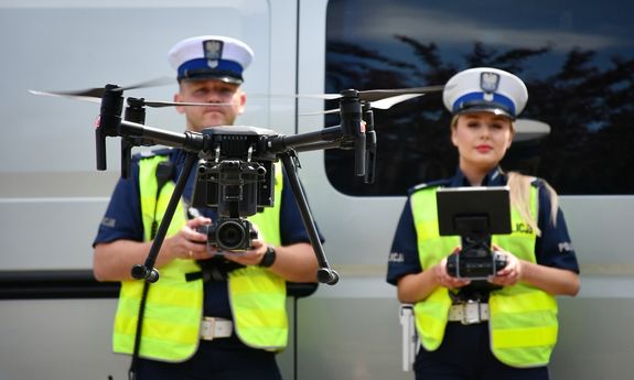 policjanci stoją przy samochodzie, przed nimi lata dron
