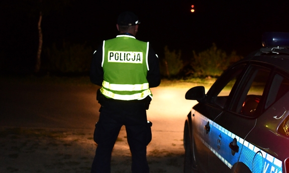 pora nocna, policjant w umundurowaniu służbowym z kamizelką odblaskową i latarką i radiowóz