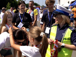 dziewczynka wiesza medal na szyi uczestniczki biegu charytatywnego, obok stoi umundurowana policjantka, która trzyma w rękach medale, w tle widać dwie osoby z identyfikatorami na szyi i uczestników pikniku
