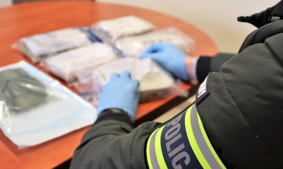 na zdjęciu na stole leżą opakowania z narkotykami, przed nimi stoi policjant, który ma na ramieniu opaskę z napisem Policja