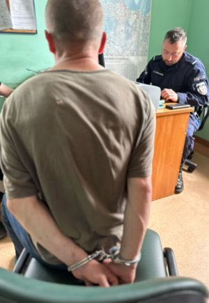 policjant siedzi przy biurku wypełniając dokumenty podczas przesłuchiwania mężczyzny