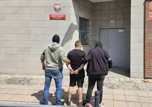 Dwaj ubrani po cywilnemu policjanci stoją z zatrzymanym mężczyzną przed aresztem śledczym. Widok z tyłu