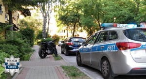 zaparkowany na chodniku motocykl, z boku widać zaparkowane przy ulicy cywilne auto, a za nim policyjny radiowóz
