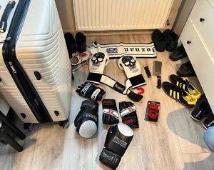 na podłodze leżą rękawice bokserskie, buty sportowe, tasak, złożony scyzoryk, po lewej stronie stoi walizka podróżna na kółkach