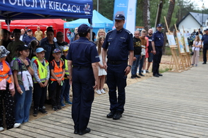 Policjantka składa meldunek oficerowi Policji, po lewej stronie zdjęcia dzieci stoją pod namiotem promocyjnym policji świętokrzyskiej