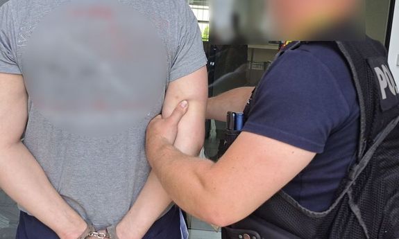 mężczyzna stojący tyłem w kajdankach oraz stojący obok niego bokiem policjant w mundurze trzymający go za ramię