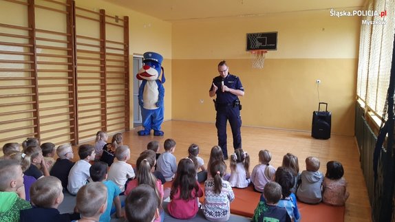Policjant w sali gimnastycznej rozmawia z grupą dzieci, po jego prawej stronie przy drzwiach stoi maskotka policyjna.