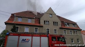 budynek mieszkalny z dachu którego wydobywa się dym, przed budynkiem widoczny samochód straży pożarnej