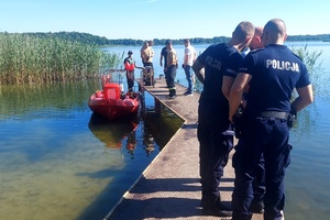 na zdjęciu umundurowani policjanci i strażacy stoją na pomoście, obok pomostu widać łódkę wykorzystywaną do poszukiwań zaginionej kobiety