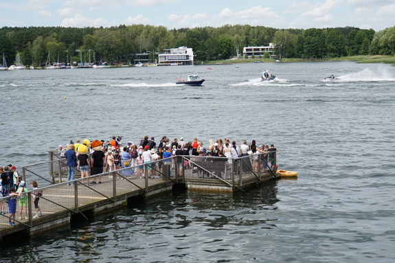 Grupa ludzi stoi na pomoście w tle pływają łodzie policyjne.