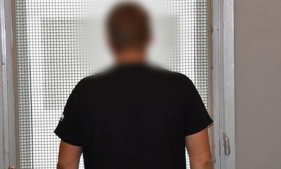 mężczyzna ubrany w czarną koszulkę, stojący tyłem do obiektywu, na tle zakratowanego okna