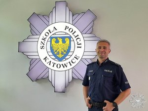 policjant stoi na tle loga Szkoły Policji w Katowicach wiszącym na białej ścianie